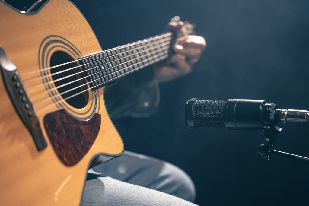 Male musician playing acoustic guitar behind microphone in recording studio.